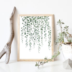 Pode incluir: Uma impressão de aquarela emoldurada de videiras verdes com folhas penduradas em um fundo branco. A moldura é de madeira marrom clara.