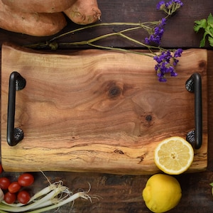 May include: A wooden serving tray with black metal handles. The tray is made of natural wood with a rustic finish. There are various food items and flowers around the tray, including red onions, green onions, sweet potatoes, lemons, and purple flowers.