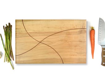 May include: A rectangular wooden cutting board with a light brown finish and two inlaid curved lines. The board is surrounded by a white background and there are various kitchen items on the surface, including a bunch of asparagus, a carrot, and a chef's knife.