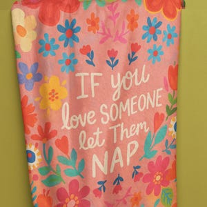 May include: A pink floral blanket with the text "IF you love SOMEONE let Them NAP". The blanket features colorful flowers in shades of red, blue, yellow, and orange. The blanket is hanging against a green wall.