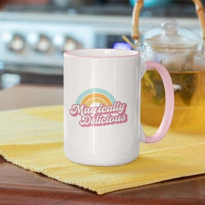 Könnte beinhalten: Weißer Keramikbecher mit rosa Henkel und Rand. Der Becher zeigt ein Regenbogen-Design und den Text "Magically Delicious" in einer Retro-Schriftart. Der Becher steht auf einer gelben Oberfläche.