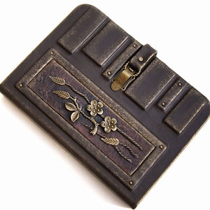 May include: A brown leather journal with a decorative metal clasp and floral design. The journal has a distressed look and is accented with gold.