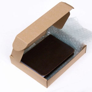 May include: A dark brown rectangular notebook with rounded corners is placed inside an open brown cardboard shipping box. A sheet of clear plastic bubble wrap is tucked along the side of the notebook within the box for protective packaging.