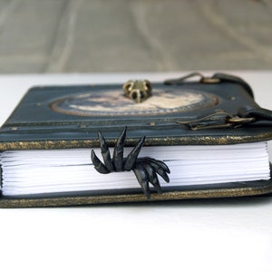 May include: A dark teal leather-bound journal with a brass clasp and decorative trim. A black claw-shaped bookmark is inserted into the pages. The journal has a vintage, gothic aesthetic.