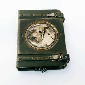 May include: Dark green leather-bound journal with a metal clasp and a decorative circular emblem. The emblem features a bird skull, a moonlit scene, and text around the edge. The journal has a distressed, antique appearance.