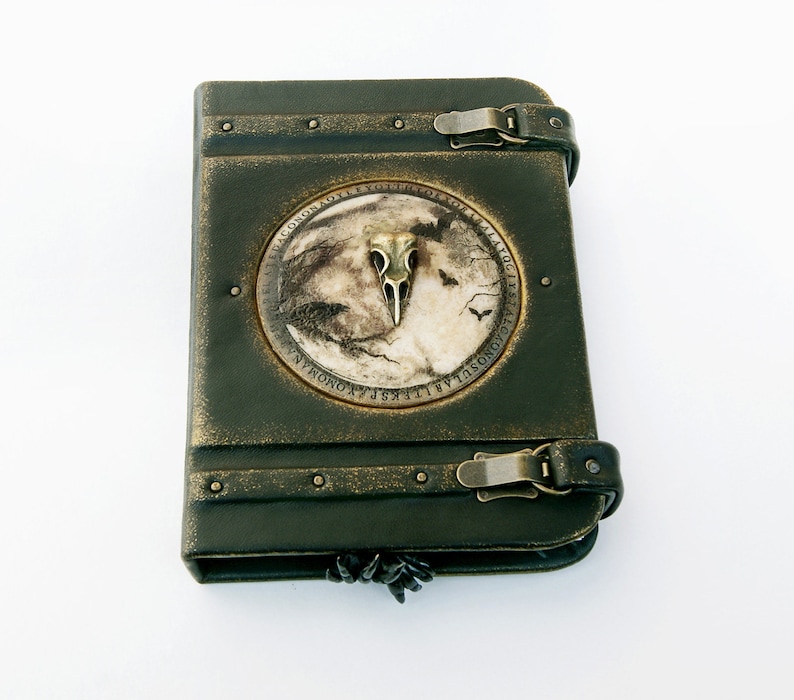 May include: Dark green leather-bound journal with a metal clasp and a decorative circular emblem. The emblem features a bird skull, a moonlit scene, and text around the edge. The journal has a distressed, antique appearance.