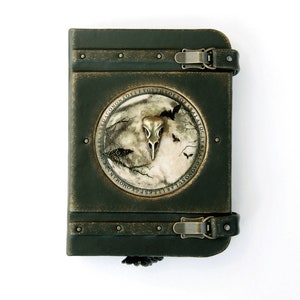 May include: A dark green, antique-style journal with a metal clasp closure. The cover features a circular emblem with a bird skull, moon, and bats. The journal is bound with dark green leather and has metal rivets.