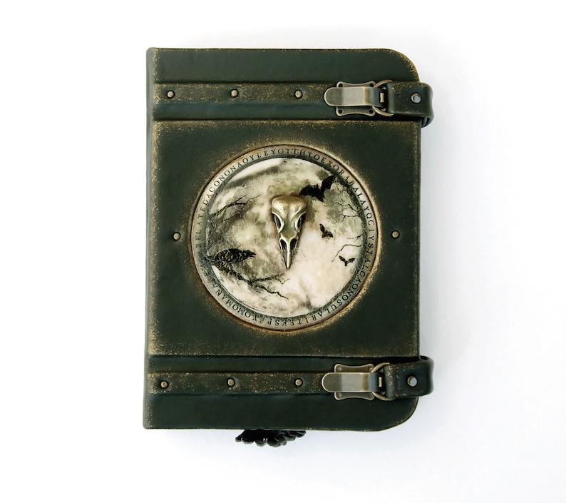May include: A dark green, antique-style journal with a metal clasp closure. The cover features a circular emblem with a bird skull, moon, and bats. The journal is bound with dark green leather and has metal rivets.