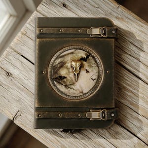 May include: A dark green box with a circular emblem depicting a bird skull, bats, and text. The box has metal details, clasps, and rivets. It rests on a weathered wooden surface.