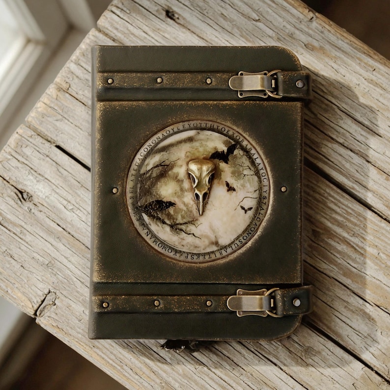 May include: A dark green box with a circular emblem depicting a bird skull, bats, and text. The box has metal details, clasps, and rivets. It rests on a weathered wooden surface.