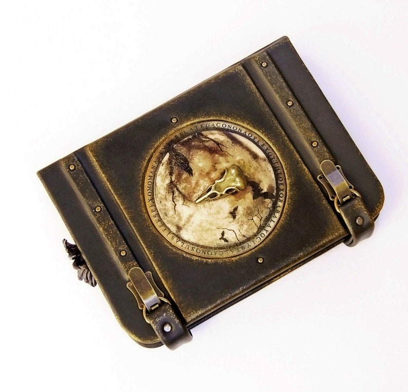 A vintage-looking book with a circular, raven scull embedded on the cover. The book features two metal clasps on its sides.