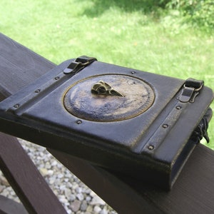 May include: A dark blue leather journal with a brass bird skull emblem on the cover. The journal features a circular design with a map-like background and is secured with two clasps. The edges of the journal have a weathered appearance, giving it a vintage look.