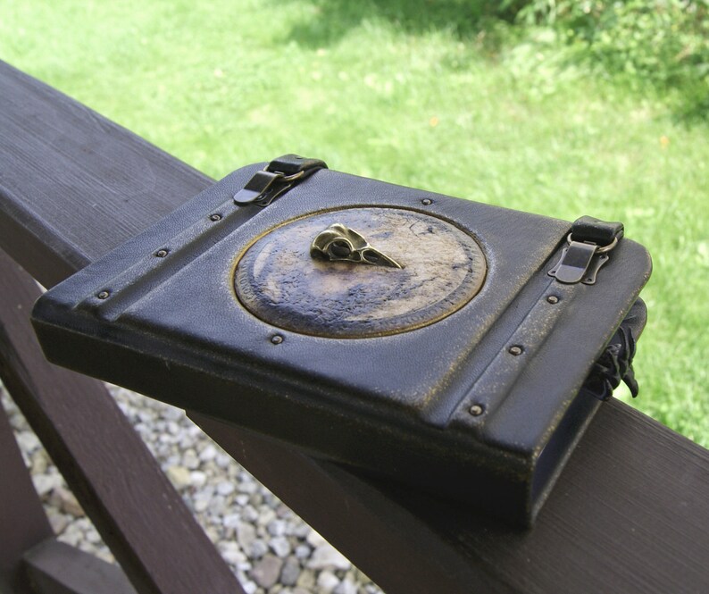 May include: A dark blue leather journal with a brass bird skull emblem on the cover. The journal features a circular design with a map-like background and is secured with two clasps. The edges of the journal have a weathered appearance, giving it a vintage look.