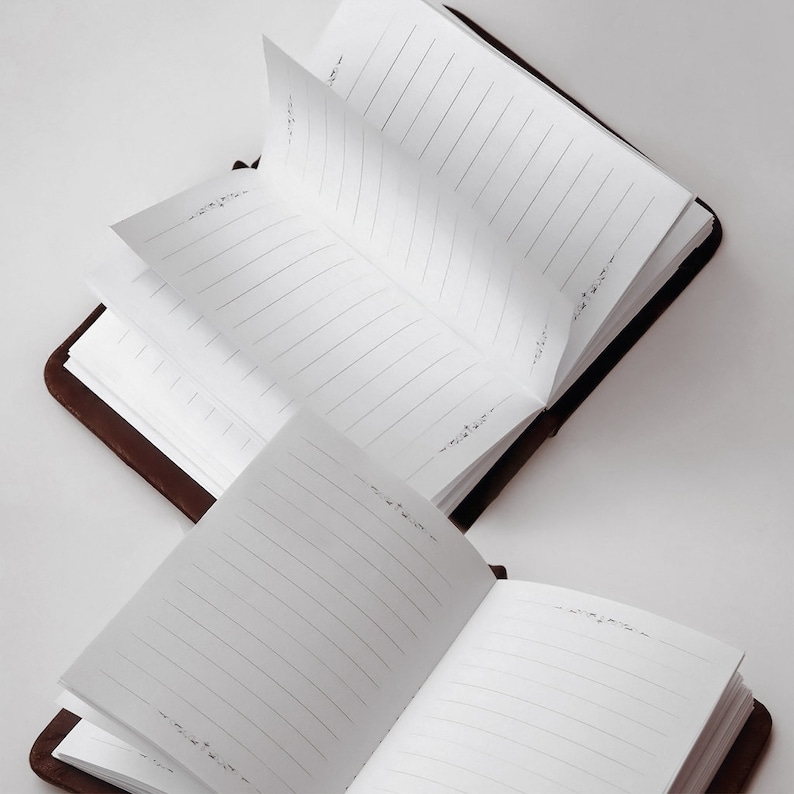 May include: Two open brown leather-bound journals with lined white pages. The pages are slightly fanned out, suggesting recent use. Delicate floral designs are printed at the top and bottom corners of the pages.