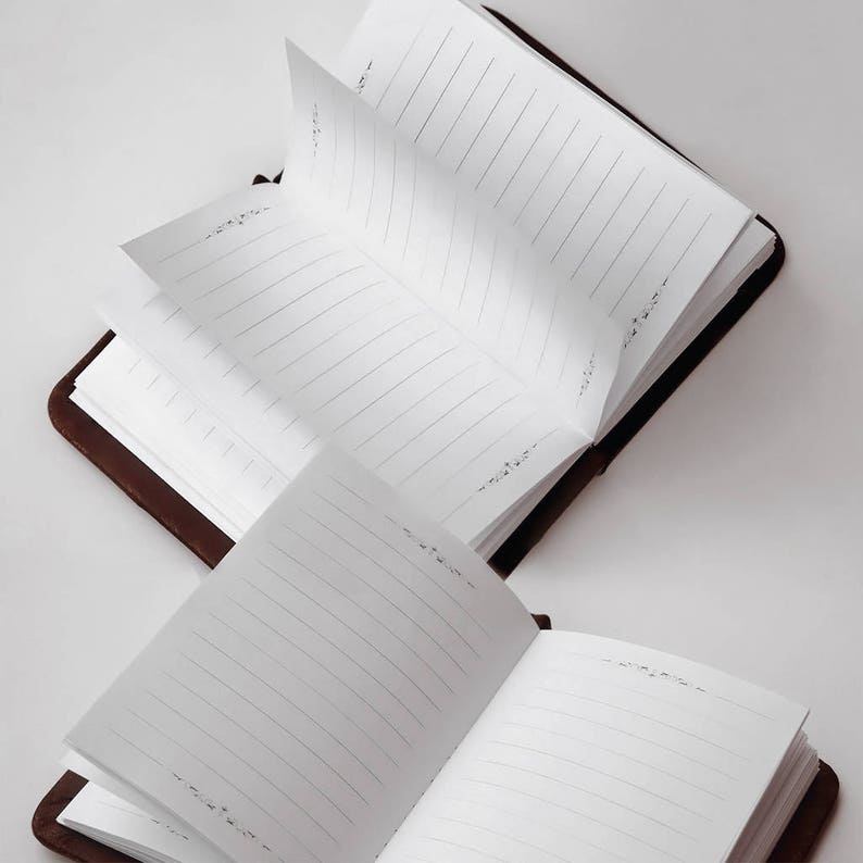 May include: Two open journals with dark brown covers and white pages. Each page features horizontal black lines for writing and small, decorative floral flourishes printed at the top and bottom margins of the paper, providing a structured layout.