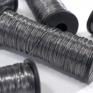 May include: Several spools of silver-colored wire are arranged on a white surface. Each spool has a black plastic core. The wire is tightly wound around the core, creating a cylindrical shape. The spools are of varying sizes.