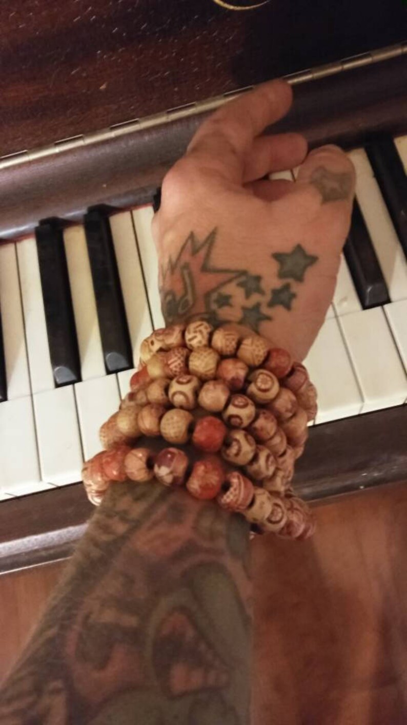 May include: A person's arm with a tattoo of stars and a lightning bolt wearing a stack of wooden bead bracelets. The bracelets are brown, red, and white.