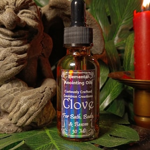 May include: A small glass bottle with a black dropper top and a label that reads "Elemental Anointing Oils Curiously Crafted Cauldron Creations Clove For Bath, Body & Besom 30 Ml".