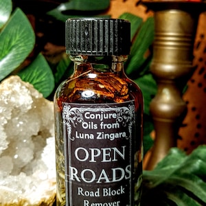 May include: A small, dark brown glass bottle with a black cap. The bottle is filled with a clear liquid and contains herbs and flowers. The label reads "Conjure Oils from Luna Zingara, Open Roads, Road Block Remover, Curiously Crafted Cauldron Creations".