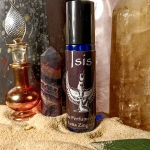 May include: A perfume bottle labeled "Isis" with a winged figure design, alongside crystals and decorative bottles. The bottle has a black cap and a dark brown label. The background includes sand and crystals.