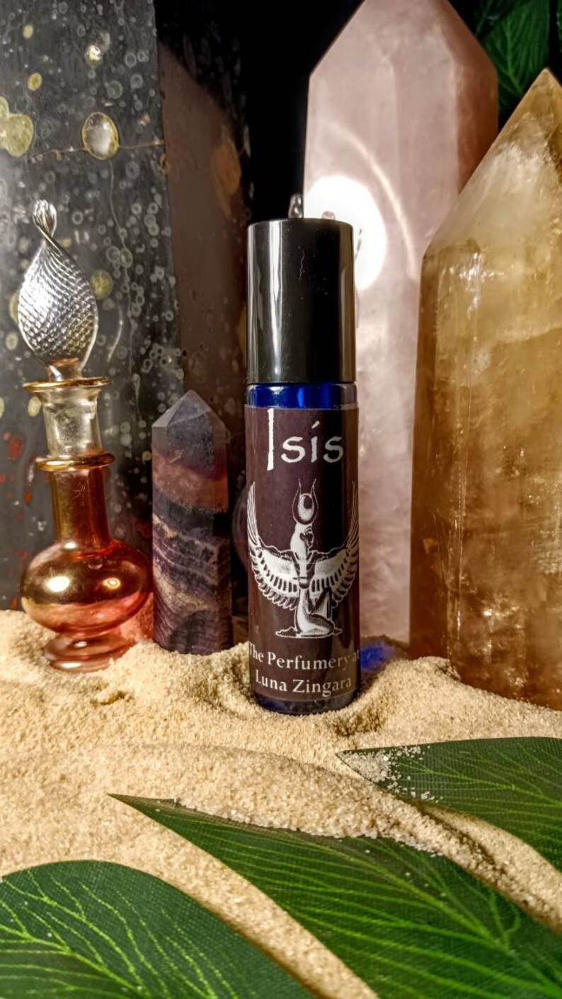 May include: A perfume bottle labeled "Isis" with a winged figure design, alongside crystals and decorative bottles. The bottle has a black cap and a dark brown label. The background includes sand and crystals.