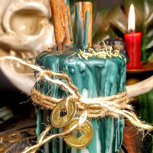 May include: A teal-colored candle with a rustic aesthetic. The candle is adorned with cinnamon sticks, gold-colored coins, and twine. A red candle burns in the background, with a crescent moon shape and greenery visible.