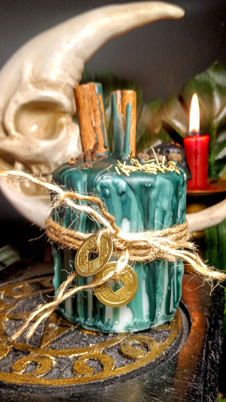 May include: A teal-colored candle with a rustic aesthetic. The candle is adorned with cinnamon sticks, gold-colored coins, and twine. A red candle burns in the background, with a crescent moon shape and greenery visible.