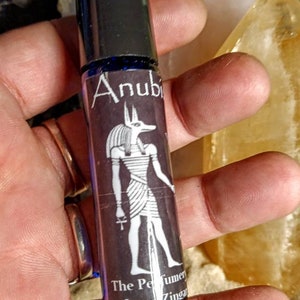 Puede incluir: Un frasco negro con rodillo y una etiqueta azul que presenta una ilustraci&oacute;n en blanco y negro de Anubis, el dios egipcio de la muerte. La etiqueta tambi&eacute;n dice "The Perfumer Luna Zingara"