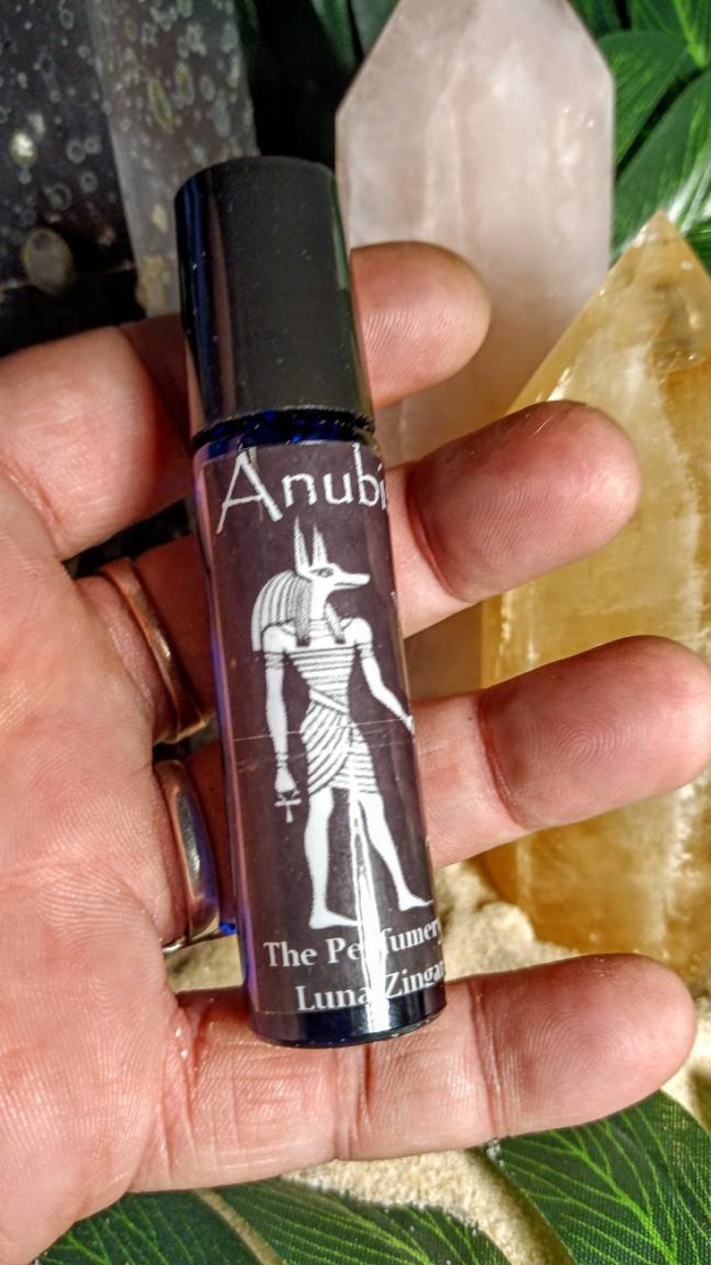 Puede incluir: Un frasco negro con rodillo y una etiqueta azul que presenta una ilustraci&oacute;n en blanco y negro de Anubis, el dios egipcio de la muerte. La etiqueta tambi&eacute;n dice "The Perfumer Luna Zingara"