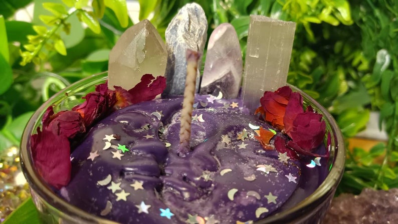May include: Purple candle with a crystal and rose petal design. The candle is in a clear glass jar and has a white wick. The candle is surrounded by crystals and rose petals.