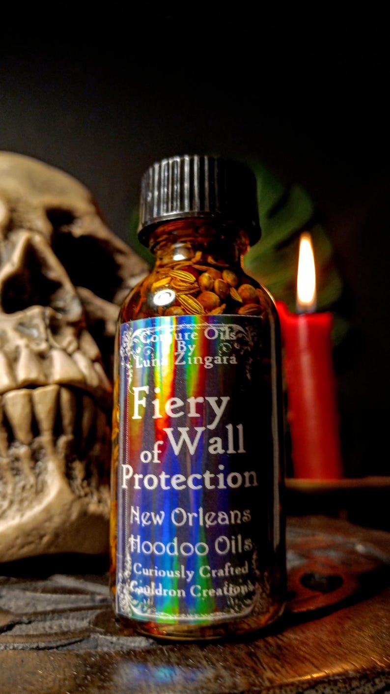 May include: A small glass bottle with a black cap filled with herbs and a label that reads "Coure Oils By Luna Zingara Fiery of Wall Protection New Orleans Hoodoo Oils Curiously Crafted Cauldron Creations".