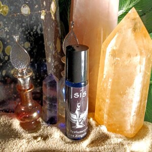 May include: A collection of crystals and perfume bottles. A roll-on perfume bottle with a black cap and the word "Isis" is centered. The background includes a large orange crystal, a pink crystal, and a dark stone.