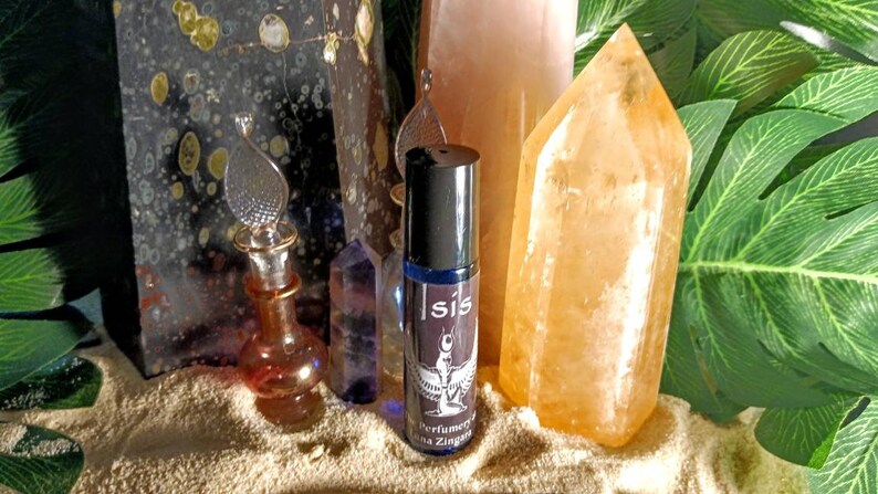 May include: A collection of crystals and perfume bottles. A roll-on perfume bottle with a black cap and the word "Isis" is centered. The background includes a large orange crystal, a pink crystal, and a dark stone.