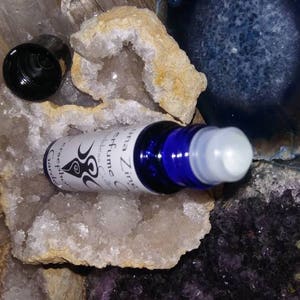 May include: A blue glass roller bottle with a white label that reads "Luna Zinna Perfume Oil" and a black and white symbol. The bottle is sitting on a white crystal.