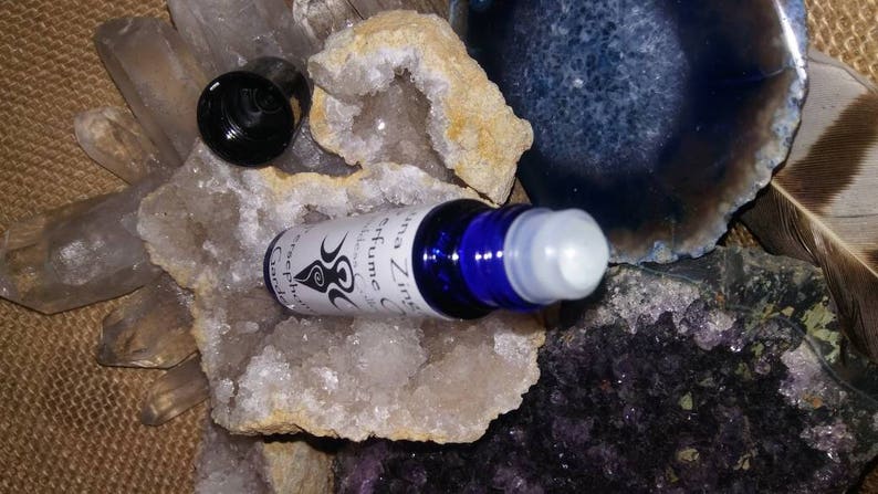 May include: A blue glass roller bottle with a white label that reads "Luna Zinna Perfume Oil" and a black and white symbol. The bottle is sitting on a white crystal.