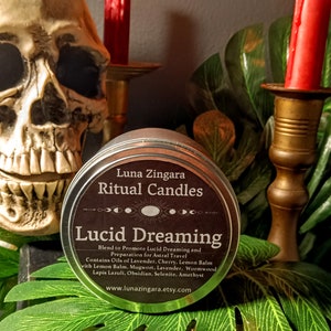May include: A black tin candle container with the text "Luna Zingara Ritual Candles" and "Lucid Dreaming" in white lettering. The candle is designed to promote lucid dreaming and astral travel. The text also lists the ingredients: Lavender, Cherry, Lemon Balm, Mugwort, Wormwood, Lapis Lazuli, Obsidian, Selenite, and Amethyst. The website address is listed at the bottom: www.lunazingara.etsy.com.