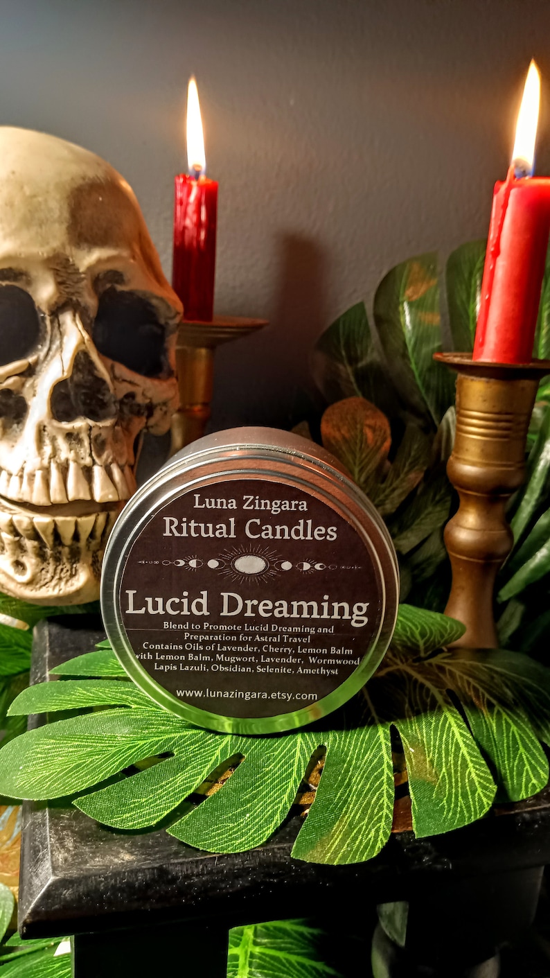 May include: A black tin candle container with the text "Luna Zingara Ritual Candles" and "Lucid Dreaming" in white lettering. The candle is designed to promote lucid dreaming and astral travel. The text also lists the ingredients: Lavender, Cherry, Lemon Balm, Mugwort, Wormwood, Lapis Lazuli, Obsidian, Selenite, and Amethyst. The website address is listed at the bottom: www.lunazingara.etsy.com.