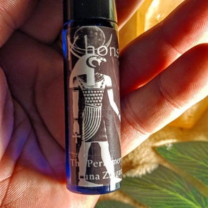 May include: A small blue glass bottle with a black cap and a label featuring a black and white illustration of an Egyptian god with a falcon head. The label also includes the text "The Perfume of Luna Za", "Khonsu", and "mer"