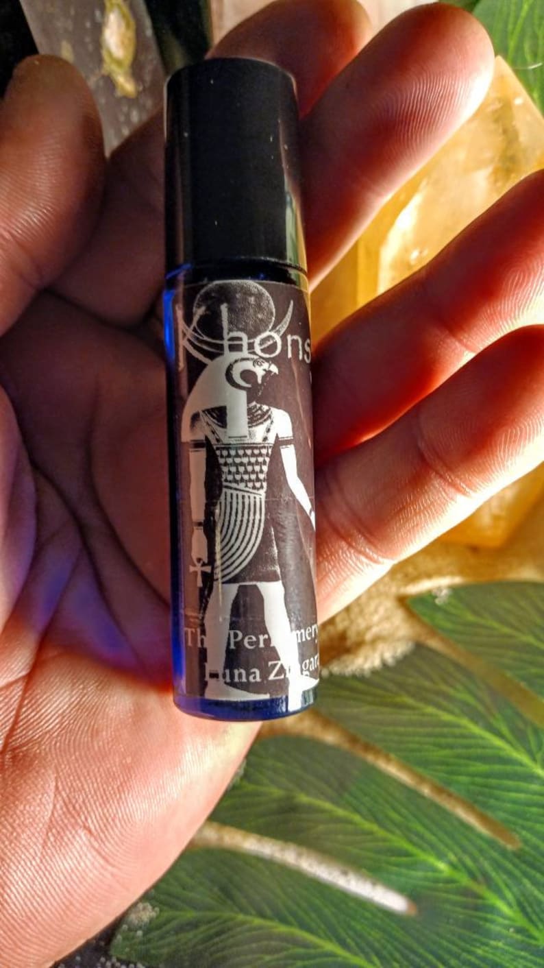 May include: A small blue glass bottle with a black cap and a label featuring a black and white illustration of an Egyptian god with a falcon head. The label also includes the text "The Perfume of Luna Za", "Khonsu", and "mer"