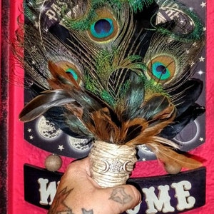 May include: A hand holds a peacock feather wand with a triple moon and pentacle charm. The wand is wrapped in twine and has brown and black feathers. The background is a red surface with a black sign that says "Welcome".