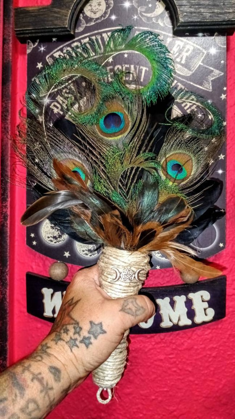 May include: A hand holds a peacock feather wand with a triple moon and pentacle charm. The wand is wrapped in twine and has brown and black feathers. The background is a red surface with a black sign that says "Welcome".