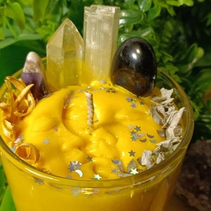 May include: A yellow candle with a clear glass jar, topped with crystals, dried herbs, and a black stone. The candle has a moon and stars design.