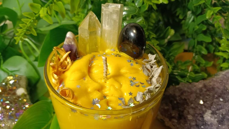 May include: A yellow candle with a clear glass jar, topped with crystals, dried herbs, and a black stone. The candle has a moon and stars design.