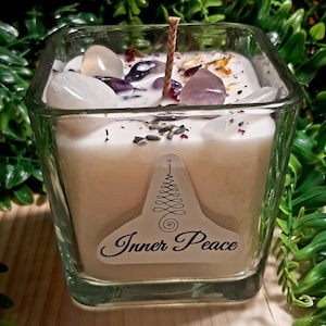 Pode incluir: Um frasco de vela quadrado de vidro com uma vela branca e um pavio de madeira. A vela &eacute; coberta com cristais transparentes e roxos e flores secas. O frasco tem um r&oacute;tulo que diz "Inner Peace" com um design de &aacute;rvore estilizado.