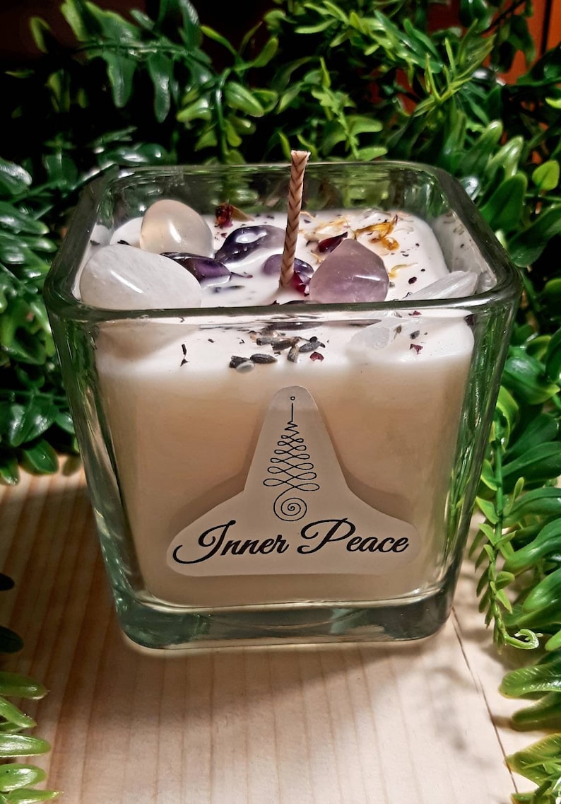 Pode incluir: Um frasco de vela quadrado de vidro com uma vela branca e um pavio de madeira. A vela &eacute; coberta com cristais transparentes e roxos e flores secas. O frasco tem um r&oacute;tulo que diz "Inner Peace" com um design de &aacute;rvore estilizado.