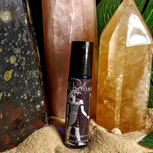 May include: A black roller bottle with a white label featuring an Egyptian god design and the text "Khonsu" and "Perfumery Alina Zungara". The bottle is sitting on a bed of sand with two crystal points behind it.