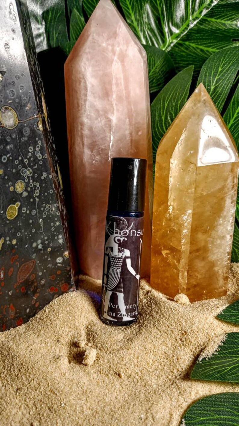 May include: A black roller bottle with a white label featuring an Egyptian god design and the text "Khonsu" and "Perfumery Alina Zungara". The bottle is sitting on a bed of sand with two crystal points behind it.