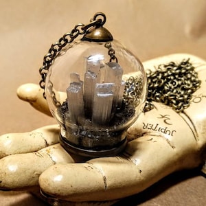 Peut inclure: Un collier avec un pendentif en forme de globe de verre et une cha&icirc;ne argent&eacute;e. Le globe contient un amas de cristaux de s&eacute;l&eacute;nite blancs sur un lit de sable noir.