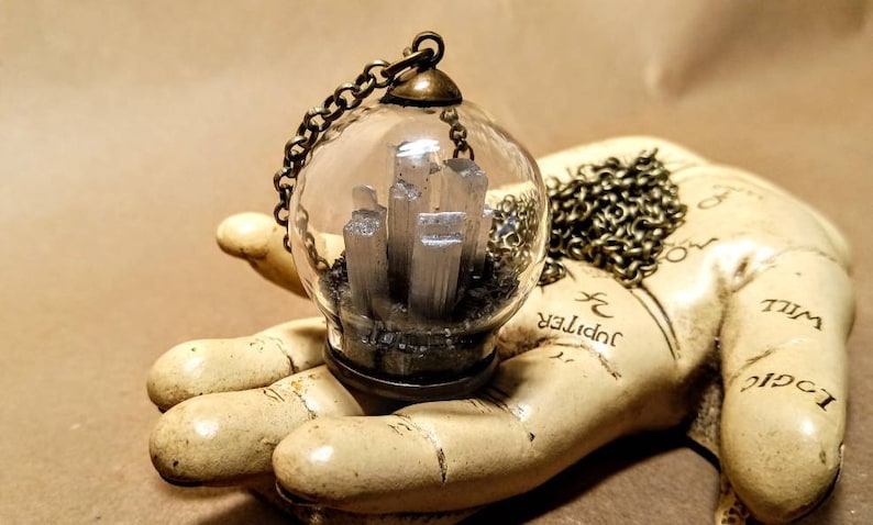 Peut inclure: Un collier avec un pendentif en forme de globe de verre et une cha&icirc;ne argent&eacute;e. Le globe contient un amas de cristaux de s&eacute;l&eacute;nite blancs sur un lit de sable noir.