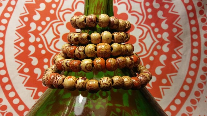 May include: A stack of five wooden bracelets with intricate patterns and brown and orange accents. The bracelets are stacked on a green bottle.